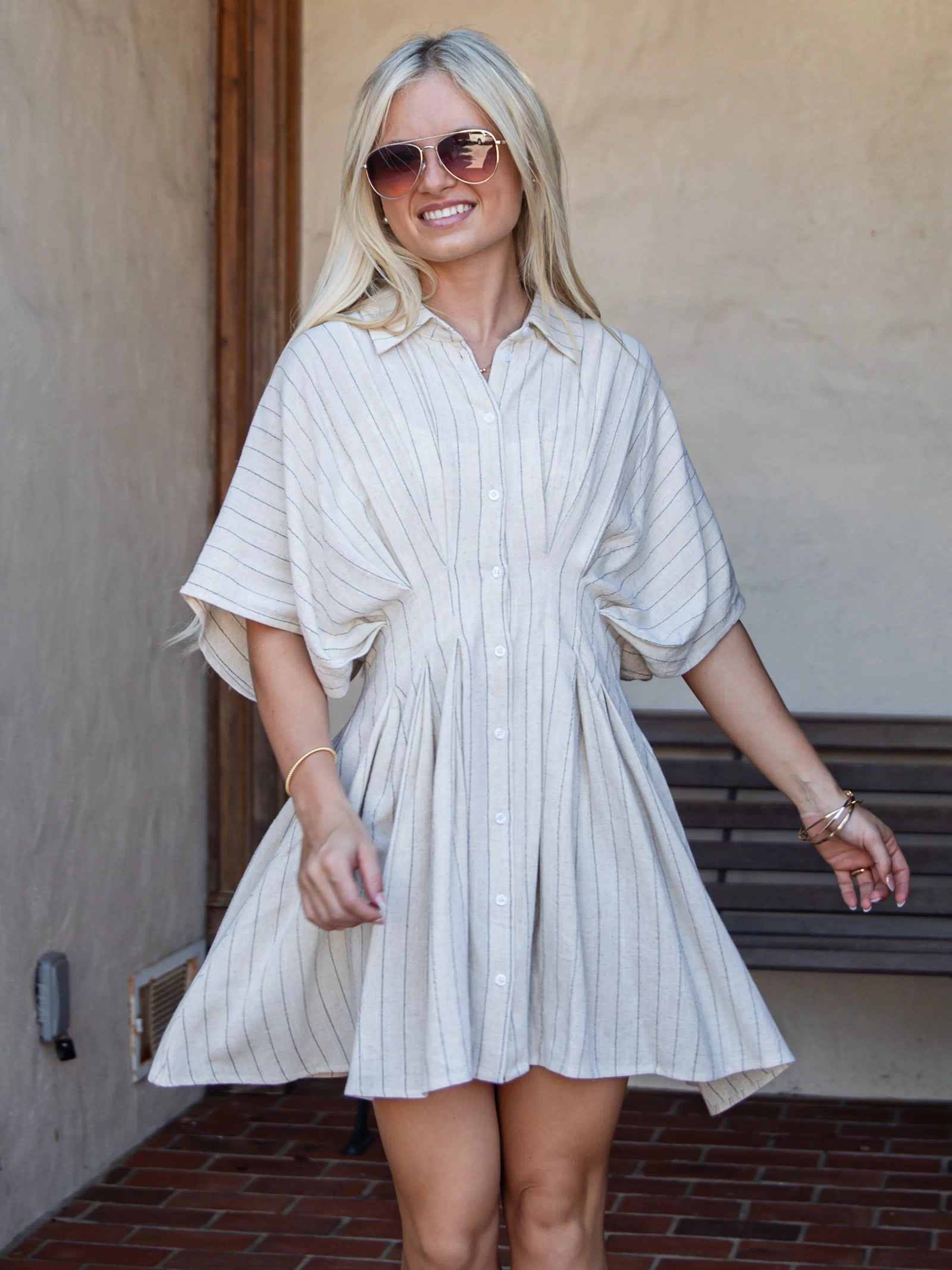A woman wearing a cream and navy blue vertically striped button-down shirt dress, standing in front of a stucco wall and brick floor.
