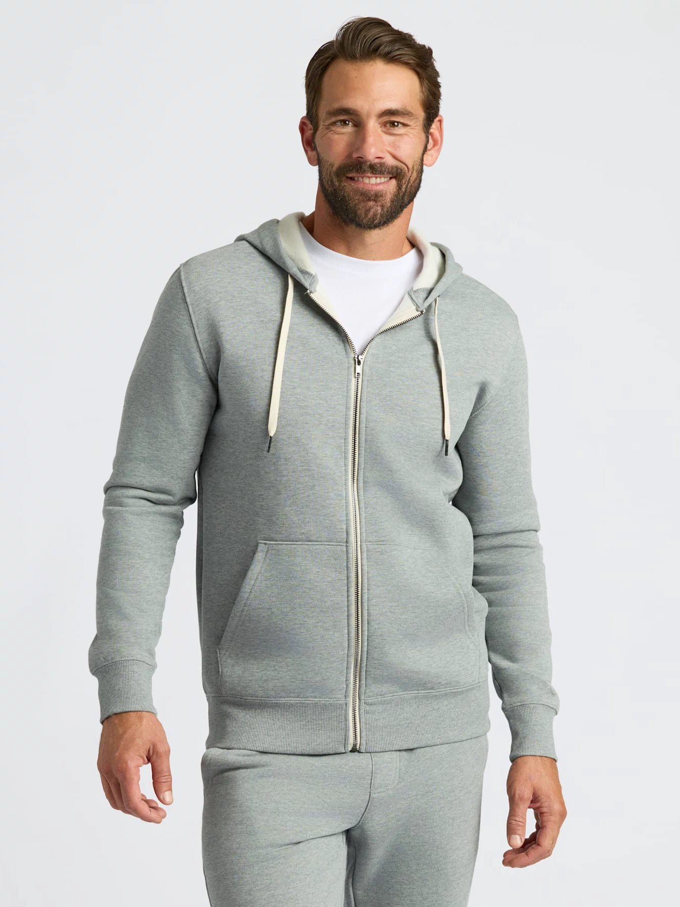 A man wearing a light gray zip-up hoodie with cream drawstrings and matching gray sweatpants, standing against a light gray studio background.