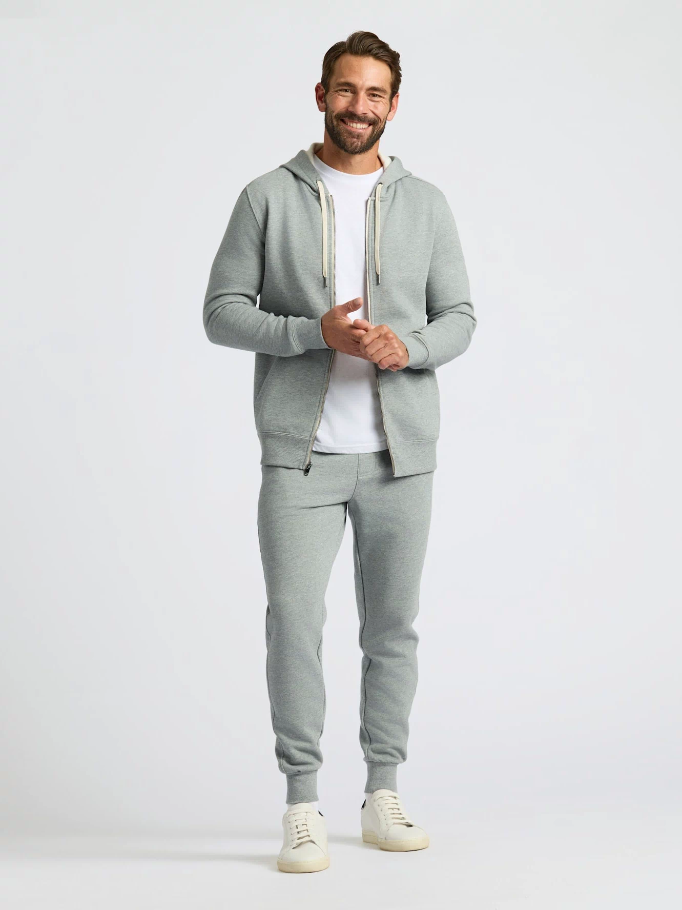 A man modeling a light gray two-piece tracksuit featuring a zip-up hoodie with white drawstrings and matching jogger pants, styled over a white t-shirt and cream sneakers against a light gray studio background.