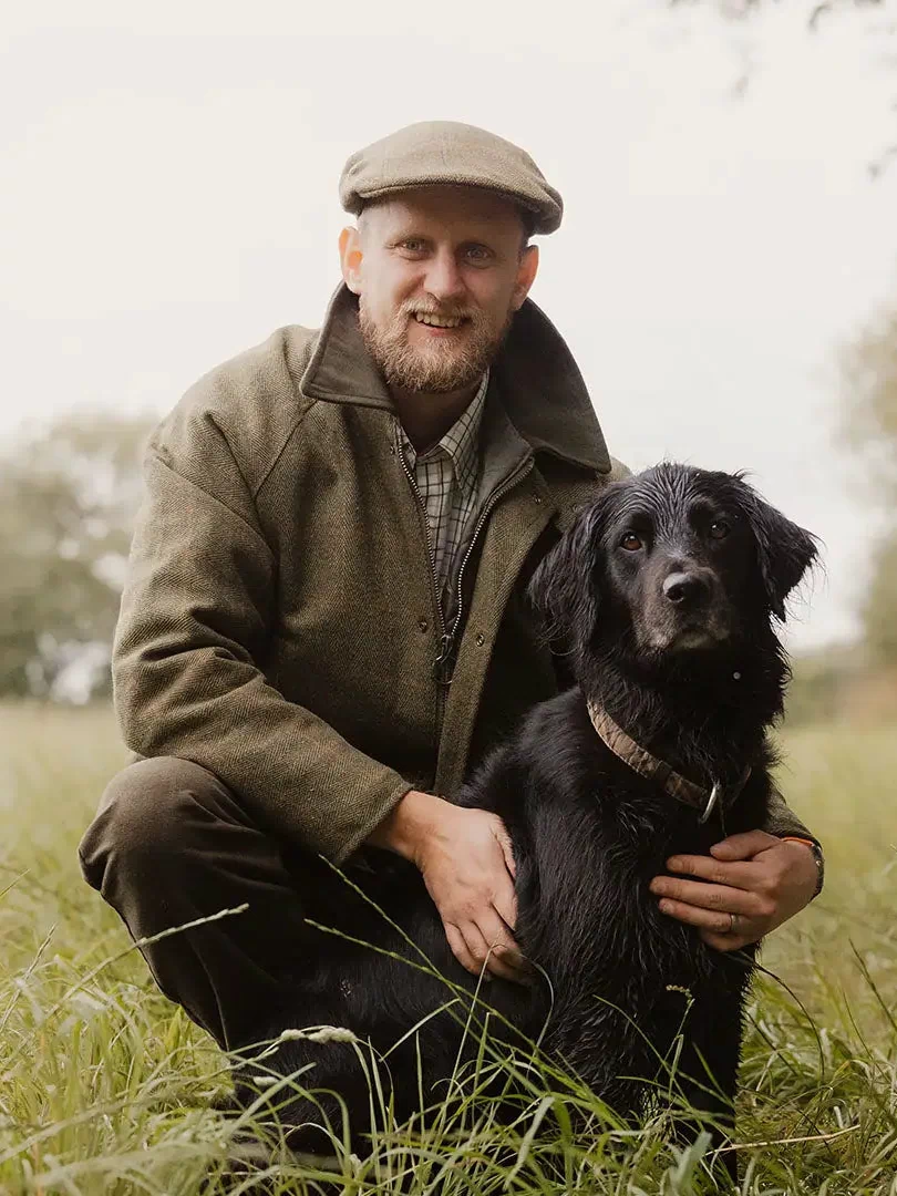 Man in a Tweed Shooting Jacket kneeling next to his black dog in a grassy field