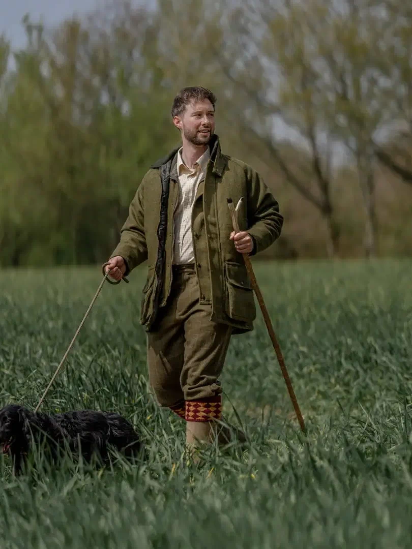 Man wearing a tweed shooting jacket walking a black dog in a grassy field