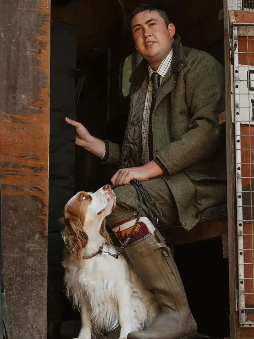 Man in a green suit with a spaniel by his feet, showcasing an English-made tweed shooting jacket