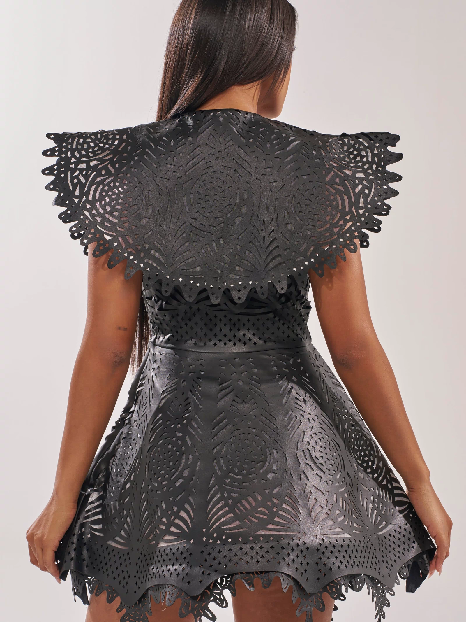 A woman with long dark hair faces away, wearing the URBAN EDGE MINI COLLARED DRESS—a black faux leather, fit-and-flare mini dress with laser-cut symmetrical patterns, scalloped shoulders, and a textured flared skirt.