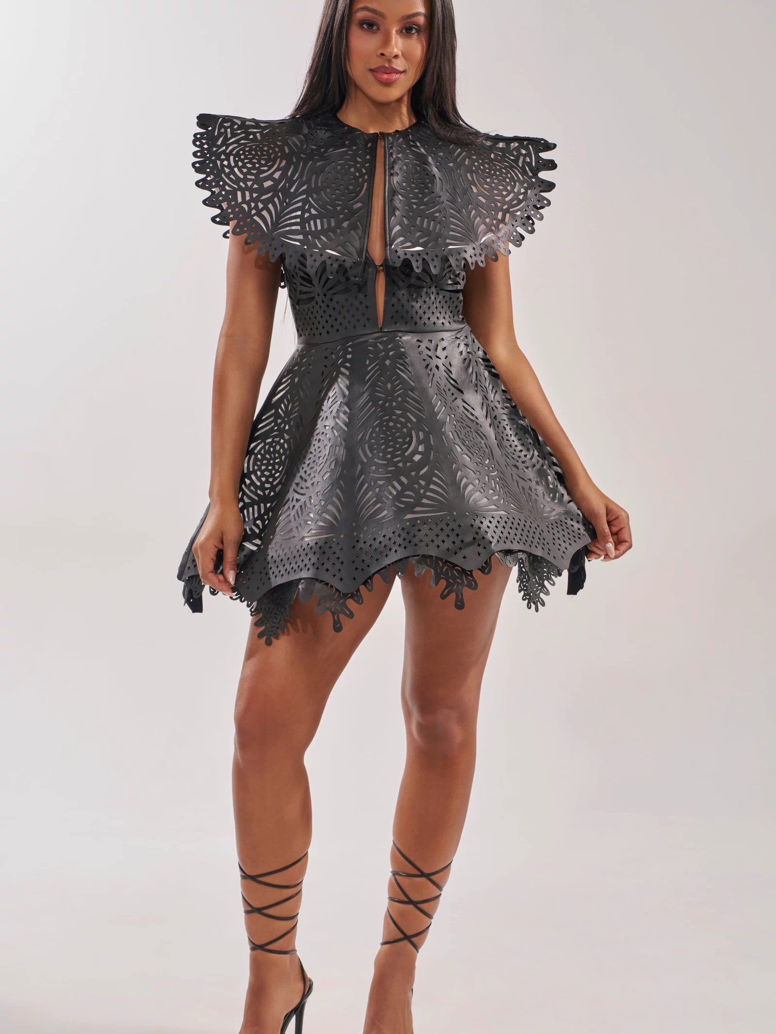 A woman poses confidently in the URBAN EDGE MINI COLLARED DRESS, featuring a black faux leather finish and a statement oversized scalloped collar. She completes the bold look with lace-up black high heels against a plain background.