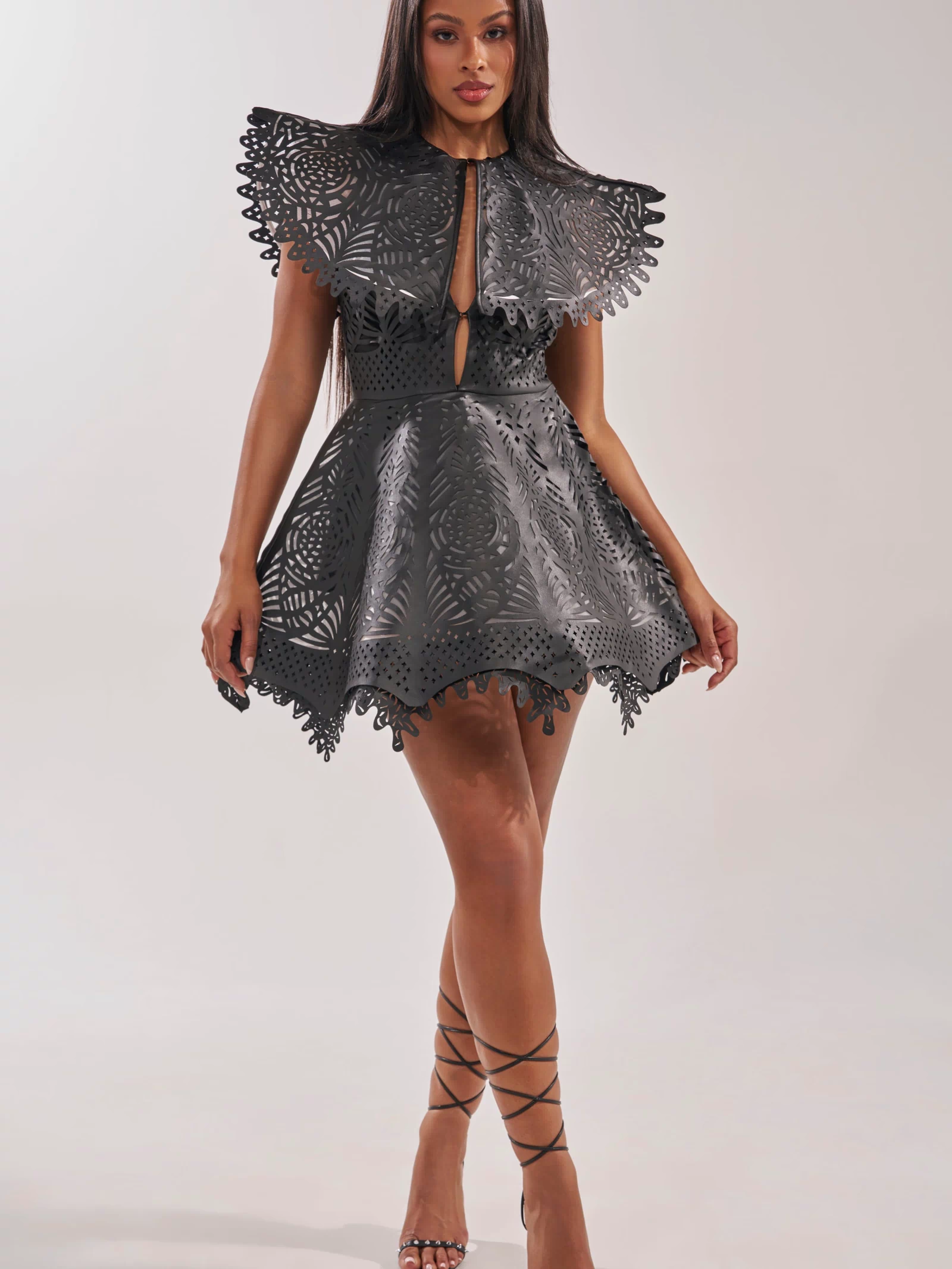 A woman wears the URBAN EDGE MINI COLLARED DRESS, a black faux leather mini with structured laser-cut patterns and a cape-style collar. She pairs it with strappy high-heeled sandals for an elegant cocktail party look.