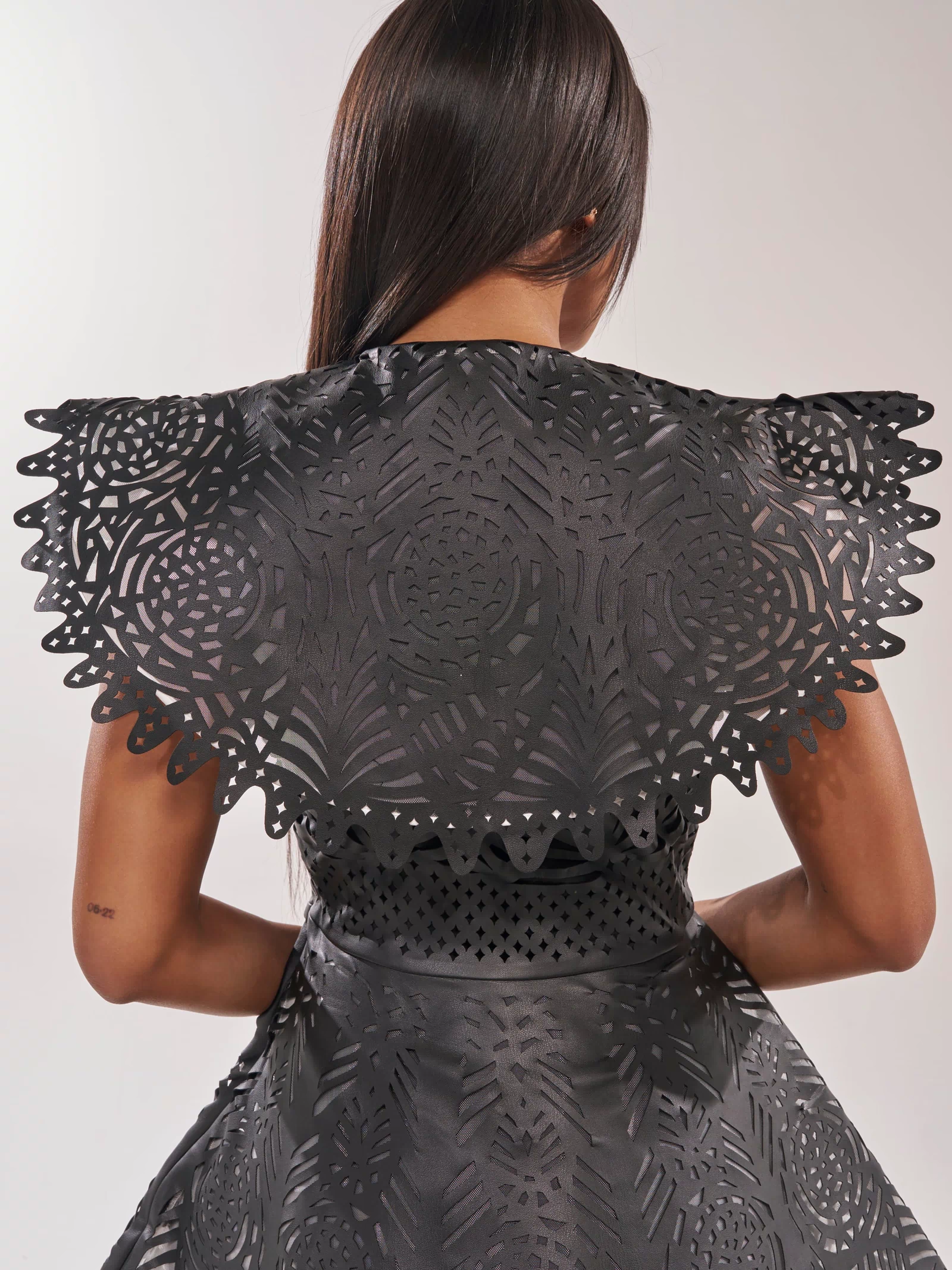 A woman with long straight hair is seen from behind in a black URBAN EDGE MINI COLLARED DRESS featuring laser-cut patterns and a dramatic collar, set against a plain light background.