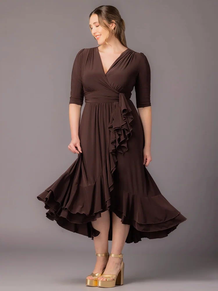 Veronica Ruffled Evening Gown in Chocolate  A curvy plus size woman stands smiling downward against a gray background in this Kiyonna maxi dress featuring a surplice V-neckline, 3/4 sleeves, wide banded waist, cascading front ruffle, and layered hi-low hem. Styled with gold platform sandals.
