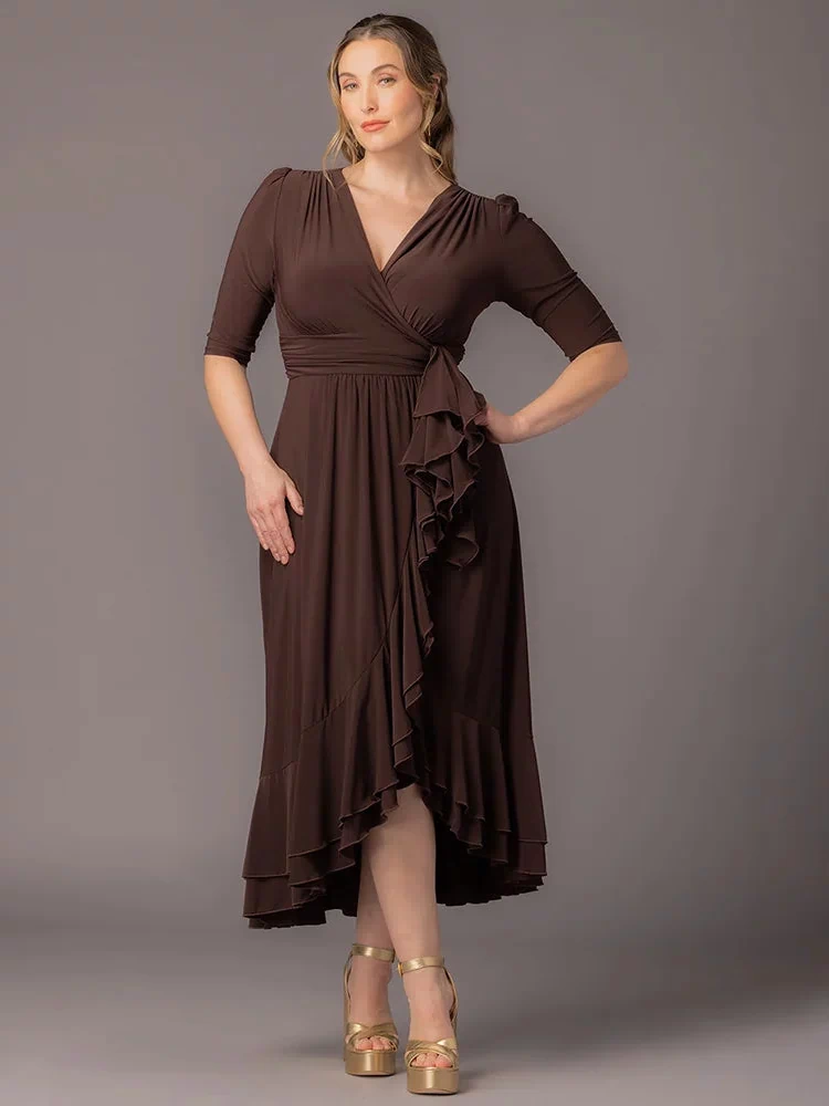 Veronica Ruffled Evening Gown in Chocolate  A curvy plus size woman poses hand on hip against a gray background in this Kiyonna maxi dress with a surplice V-neckline, 3/4 sleeves, wide banded waist, cascading front ruffle, and layered hi-low hem. Styled with gold platform sandals.