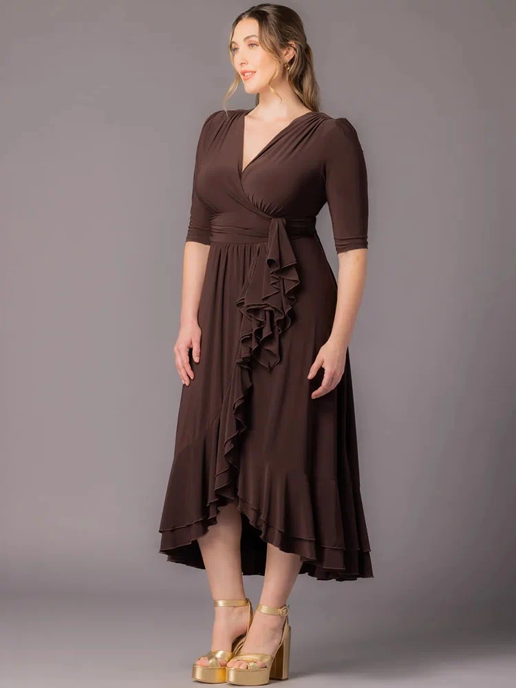Veronica Ruffled Evening Gown in Chocolate  A curvy plus size woman stands at a slight angle against a gray background in this Kiyonna maxi dress with a surplice V-neckline, 3/4 sleeves, wide banded waist, cascading front ruffle, and layered hi-low hem. Styled with gold platform sandals.