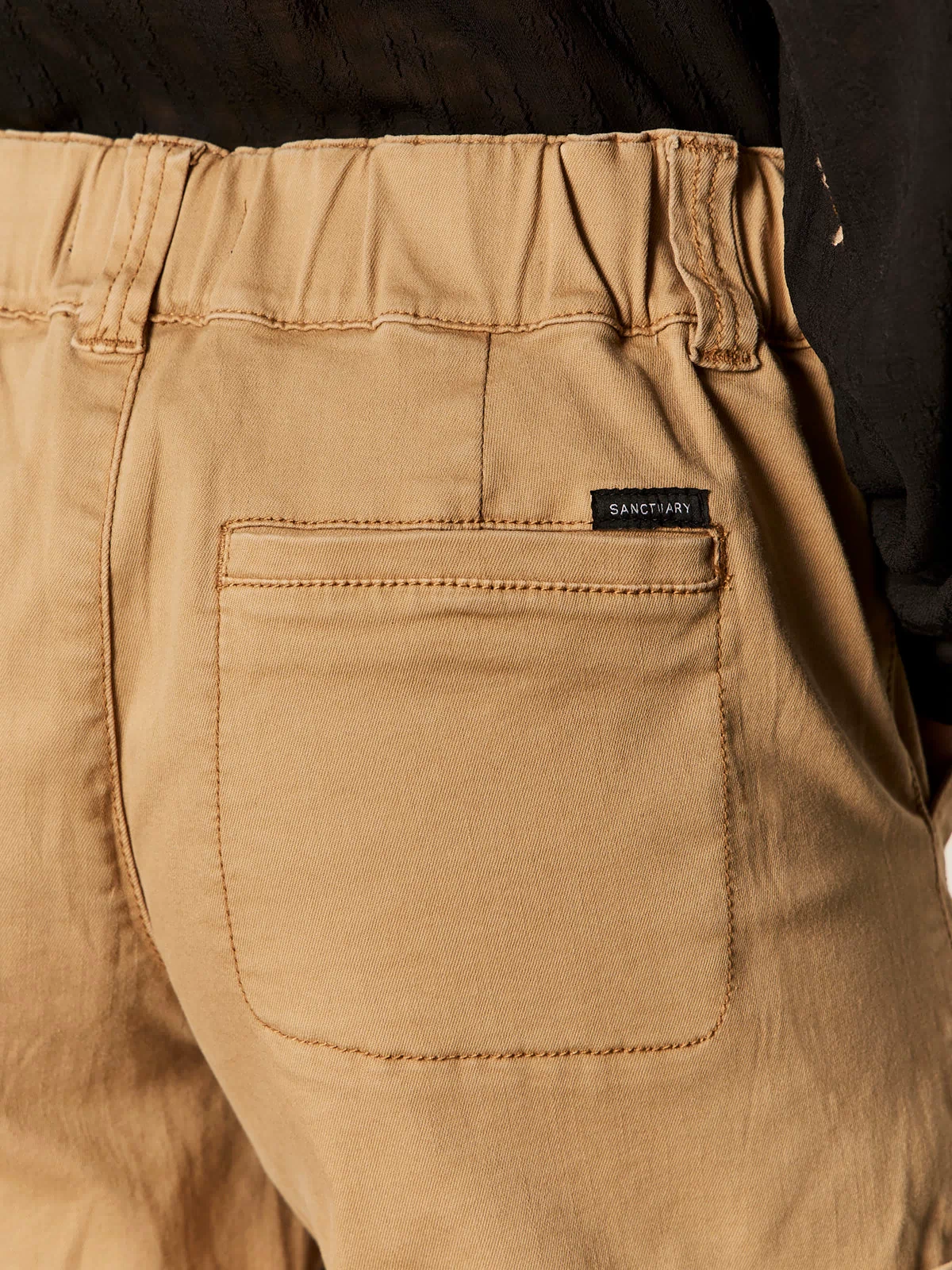 Close-up of the back pocket of Sanctuary Clothing's wander on short washed cartouche, featuring an elastic waistband and a black "Sanctuary" tag. Part of a black shirt is also visible.