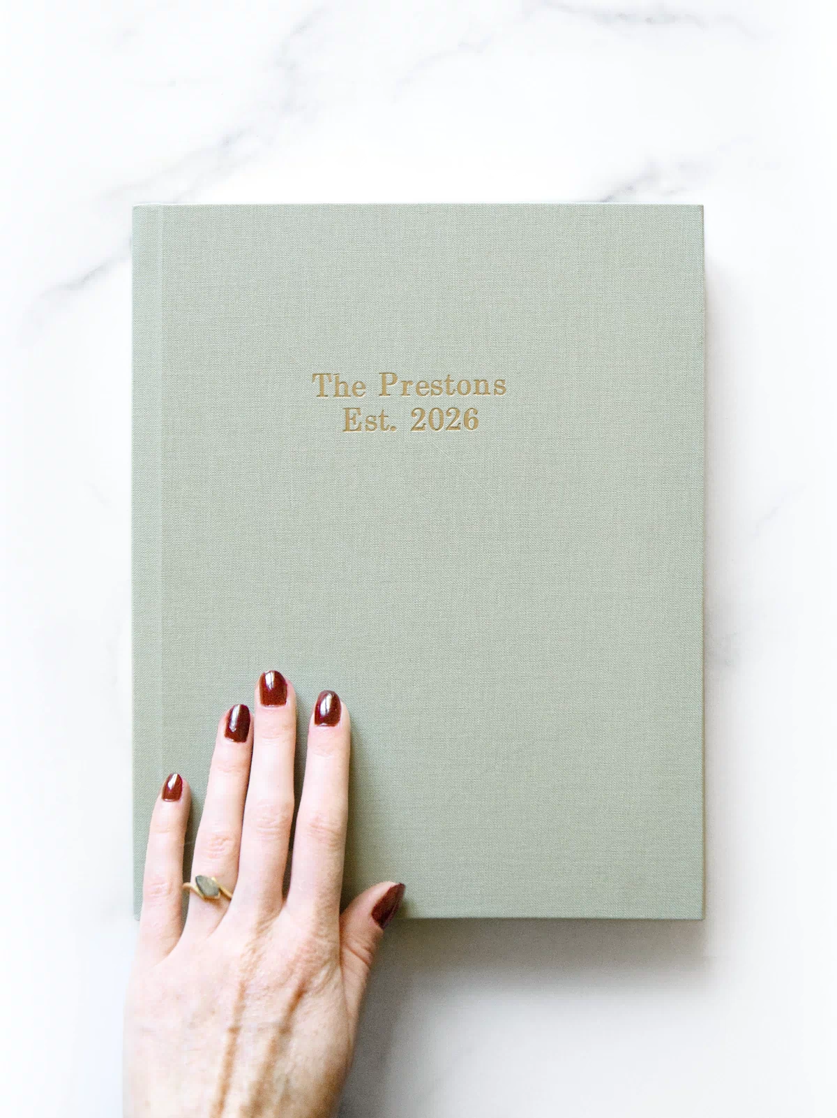 Light green book with 'The Prestons Est. 2026' text, held by a hand with red nail polish on a white background
