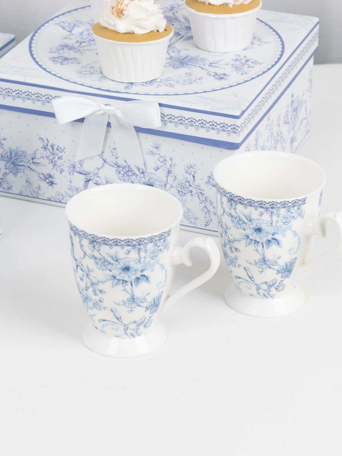 Blue floral porcelain mugs beside decorative gift boxes and cupcakes.