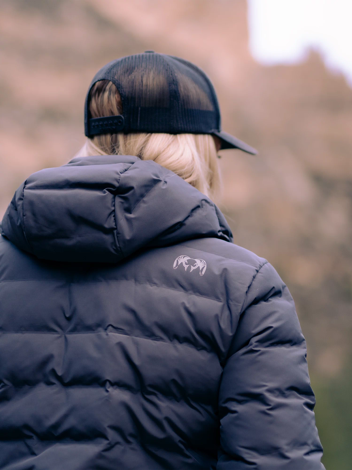 Back of woman wearing Elements Hooded Jacket in Gunmetal with KUIU Ram Logo on upper back right shoulder.