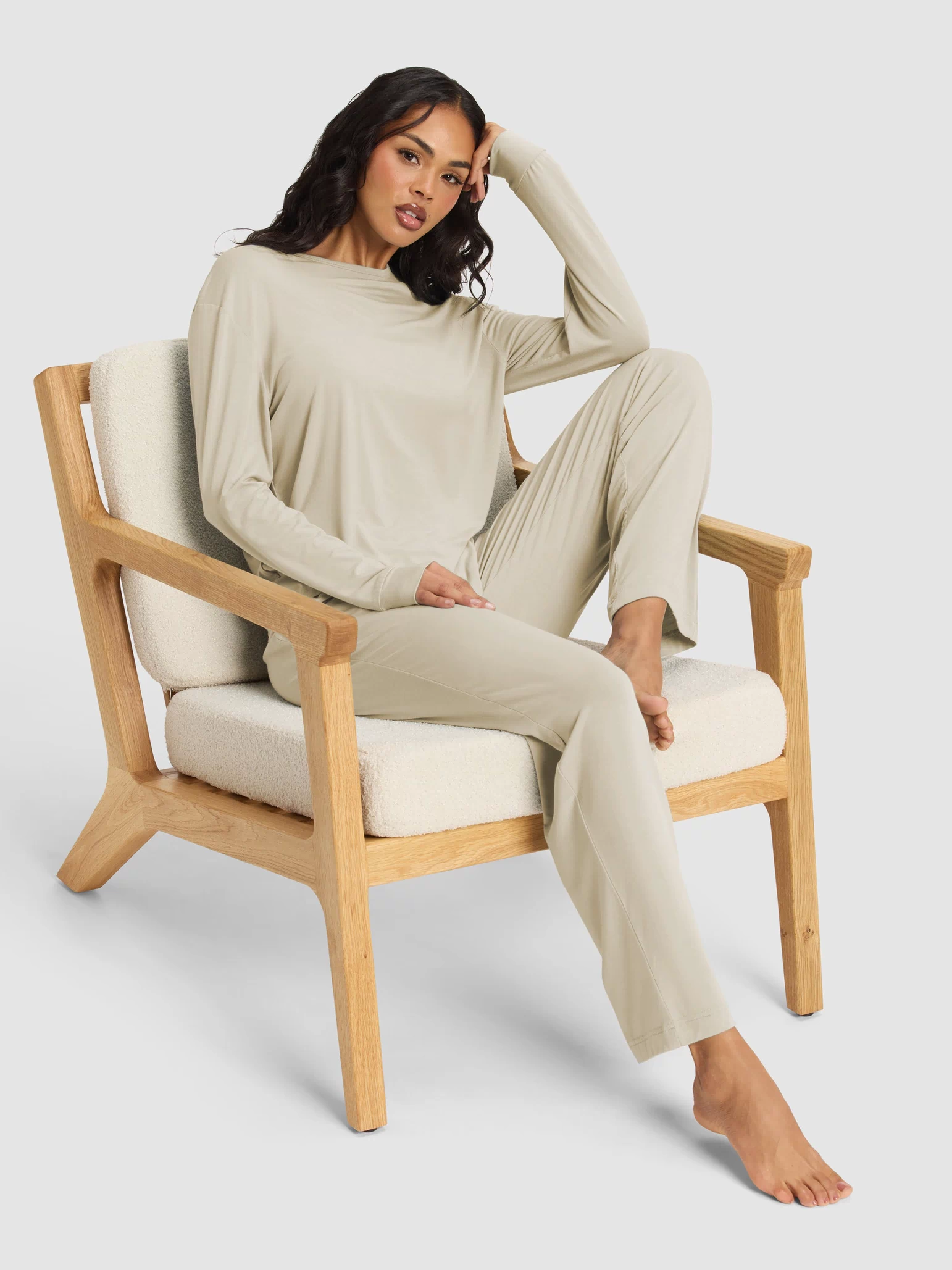 A woman relaxes barefoot in a wooden armchair with cream cushions, wearing the Cozy Earth Women's Loews Hotel Lounge Set—a light beige long-sleeve top and matching pants—with one arm resting on her head.