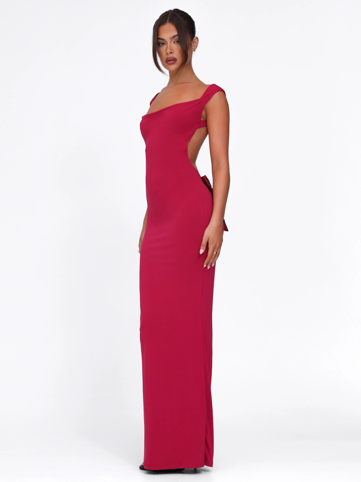 A woman poses against a plain white background wearing the TIGERMIST ZITA DRESS in deep red, a fitted, floor-length style with a bodycon silhouette, sleeveless design, open back, and bow detail.